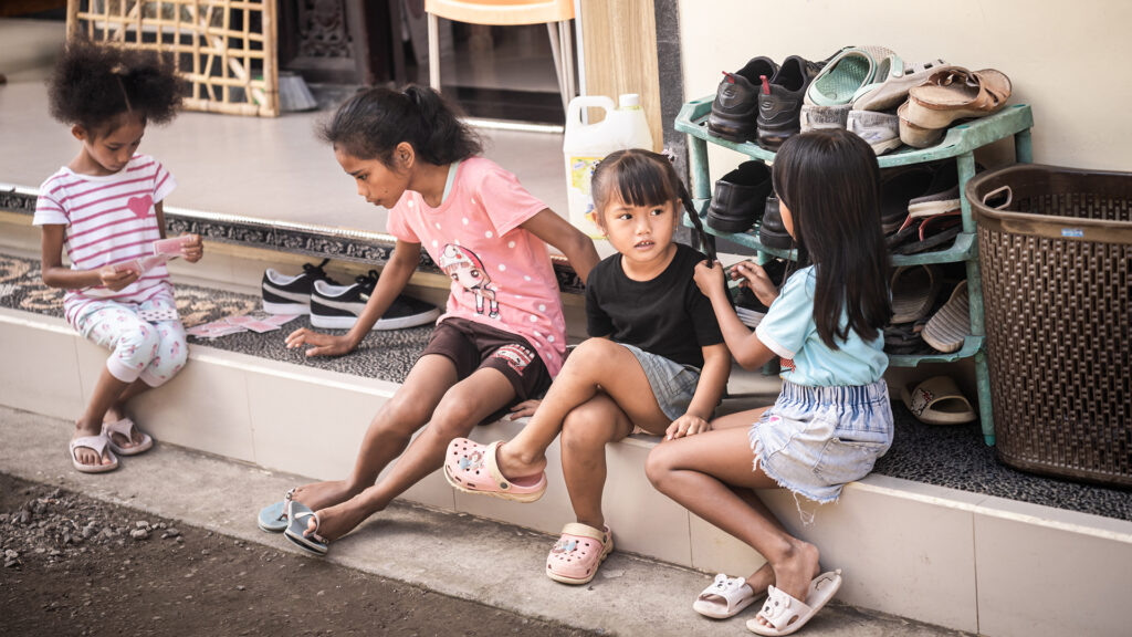 Four children are sitting on a step outside a house, with two of them playing with cards and engaging with each other. There's a rack of shoes beside them.